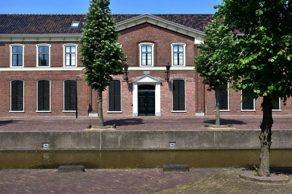 Een breed bakstenen pand met luiken en een zwarte voordeur aan een gracht in Franeker, met bomen op de kade